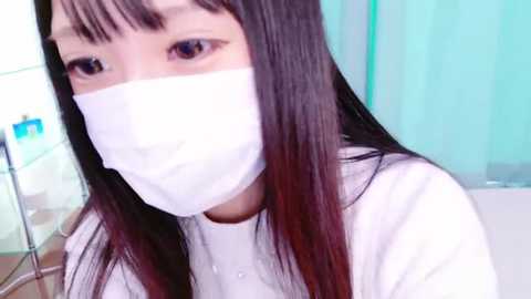 Media: A video of an East Asian woman with long, straight black hair and bangs, wearing a white surgical mask. She is in a sterile hospital room with a green wall and medical equipment in the background.