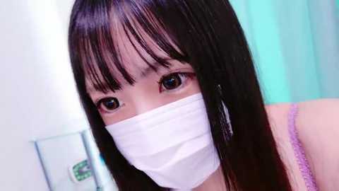 Media: Video of a young East Asian woman with straight black hair and bangs, wearing a white surgical mask, a purple spaghetti strap top, and large silver hoop earrings. Background features a light green curtain.