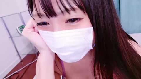 Media: A close-up video of an East Asian woman with long black hair and bangs, wearing a white surgical mask, lying on a bed in a dimly lit room.