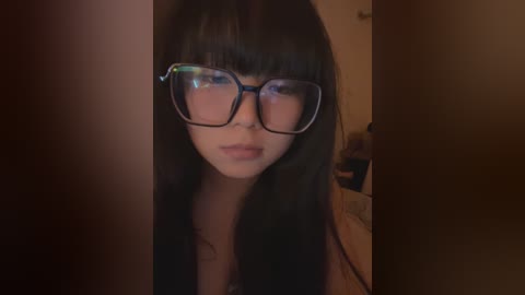 Media: Video of a young Asian woman with long black hair, wearing large black-framed glasses, standing in a dimly lit room with a blurred background.