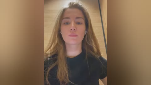 A video of a young Caucasian woman with long, straight blonde hair, wearing a black t-shirt, standing in a narrow, beige hallway with wooden floors.