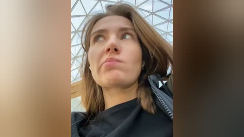 A video of a young woman with light skin and shoulder-length brown hair, wearing a black jacket, looking upward in a glass-encased, geometrically-patterned atrium.