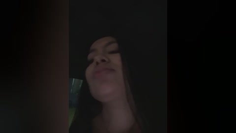 Media: A dimly lit video of a young woman with long, dark hair and light skin, smiling slightly, captured from an angle that distorts her face. The background is dark and indistinct.