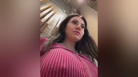 Media: A video of a young woman with long, dark hair, wearing a pink, ribbed knit top, standing in a modern, industrial-style room with exposed wooden beams and concrete walls.