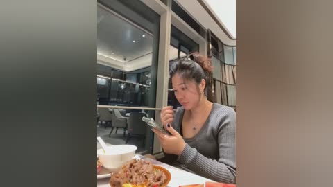 Media: Video of an Asian woman with dark hair in a bun, wearing a grey long-sleeve top, eating noodles with chopsticks at a modern restaurant table.