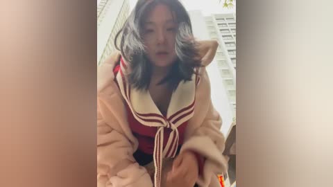 Media: Video of an Asian woman with shoulder-length black hair, wearing a red and white sailor-style top, and a beige fur coat, standing in front of high-rise buildings.