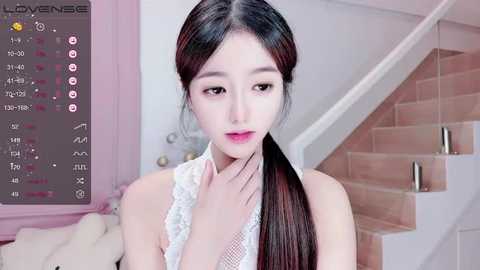 Video of an East Asian woman with long, straight black hair, fair skin, wearing a white lace sleeveless top, standing indoors near a staircase.