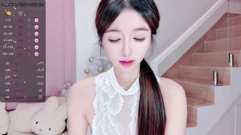 Media: Video of an East Asian woman with long black hair tied in a low ponytail, wearing a white lace sleeveless top. Background includes a pink wall, a teddy bear, and a brick staircase.