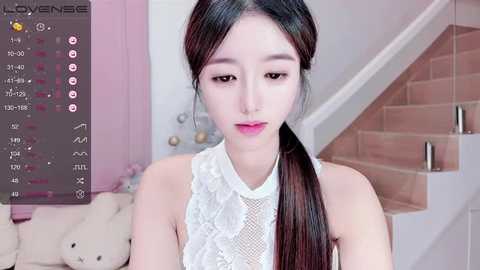 Media: A video of an East Asian woman with long, straight black hair tied in a low ponytail, wearing a white lace top, standing in a modern, well-lit room with a beige staircase and pink wall.