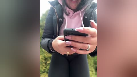 Media: Video of a woman with light skin tone, dark hair, wearing a black puffer jacket over a pink hoodie and black leggings, holding a black smartphone. Background features blurred greenery.