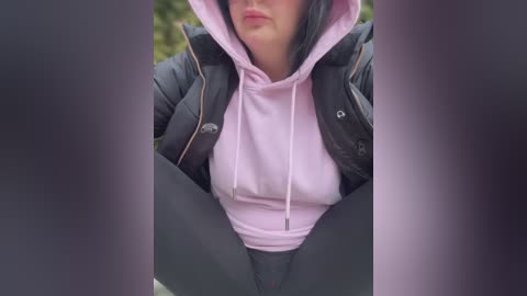 Media: Video of a woman in a pink hoodie, dark leggings, and puffer jacket, sitting with legs spread, outdoors. Her lips are slightly pursed, and her face is partially obscured by the jacket hood.