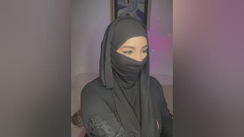 Media: Video of a light-skinned woman with a fair complexion wearing a black hijab and long-sleeved black dress, standing indoors with a blurred, colorful background.