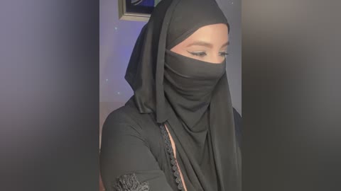 Media: Video of a woman with light skin wearing a black hijab and niqab, her face partially covered. She is indoors, with a framed picture and a blue wall visible in the background.