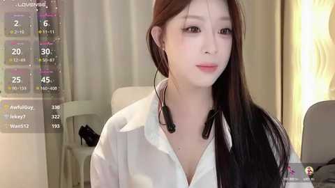 Media: Video of an East Asian woman with long, straight brown hair, wearing a white blouse and headphones, sitting in a modern, light-colored room.