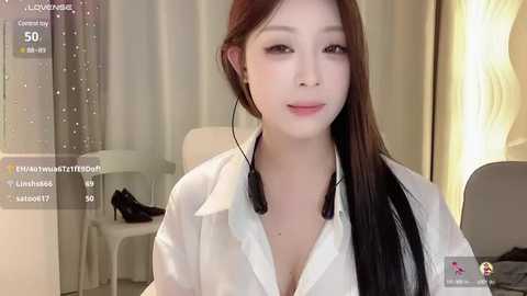 Media: Video of a young Asian woman with fair skin and long black hair, wearing a white blouse, in a modern, softly lit room with a lamp and beige curtains.