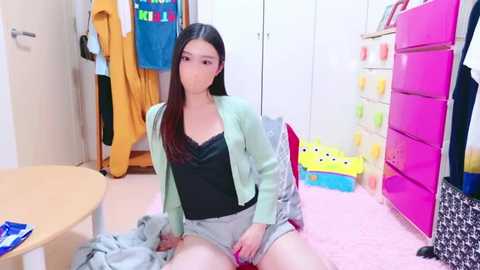 Media: Video of an Asian woman with long black hair, wearing a green cardigan and grey shorts, sitting on a pink carpet in a cluttered bedroom with colorful toys, clothes, and a dresser.