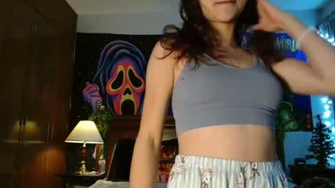 Media: Video of a young woman with long brown hair in a gray sports bra and striped pajama shorts, standing indoors. Background features a colorful, stylized painting and a wooden dresser with a lamp.