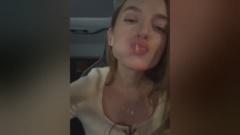 Media: Video of a young woman with fair skin and blonde hair, puckering her lips and blowing a kiss. She wears a white top, with a blurred background featuring a dark room and a white object.