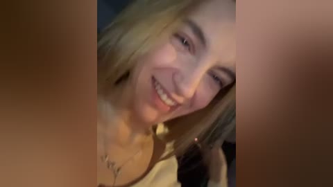 Media: Video of a young woman with light skin and blonde hair, smiling, with blurry faces in the foreground. She's wearing a white top.