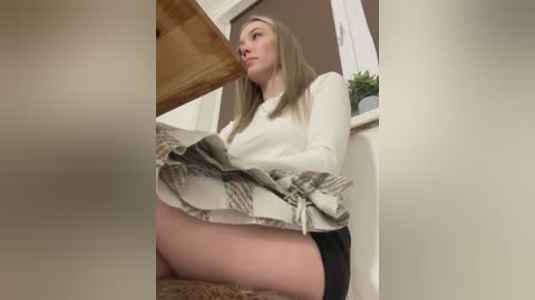 Media: Video of a young Caucasian woman with straight blonde hair, sitting on a beige carpet, wearing a white long-sleeve shirt and a plaid skirt, looking contemplative.