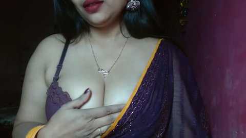 Media: Video of a woman with long black hair, wearing a purple saree with golden trim, revealing ample cleavage. She has fair skin and wears large, ornate earrings.