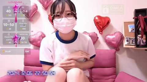 Media: A video captures a young Asian woman with glasses, wearing a white t-shirt and pink mask, sitting on a pink couch. The background features heart-shaped pink decorations, a clock, and a framed photo.