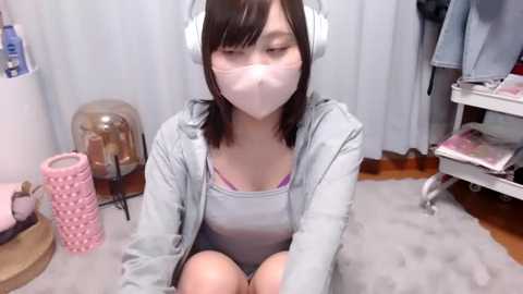 Media: Video of an Asian woman with short black hair wearing a face mask, headphones, and a light gray hoodie, sitting on a gray carpet in a cluttered room with pink and white decor.