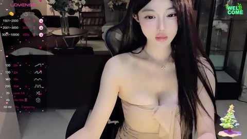 Media: A young East Asian woman with long black hair, fair skin, and a slender physique, wearing a beige tube top, sits on a black office chair in a modern living room.