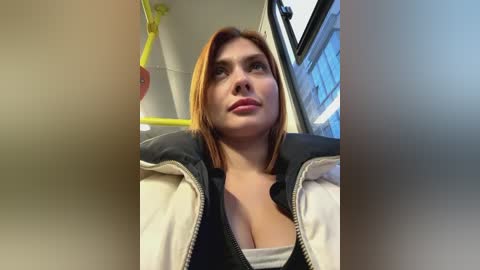 Media: Video of a young woman with fair skin and shoulder-length auburn hair, wearing a black top and white jacket, standing on a bus. Background includes yellow handrails and a window.