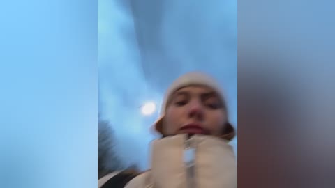 Media: Video of a young woman with light skin, wearing a beige puffy jacket and matching beanie, gazing downward with a contemplative expression. Background shows a snowy landscape with blurred trees and a bright moon in the sky.