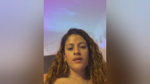 Media: Video of a young woman with curly brown hair, wearing a pink top, looking into the camera with a neutral expression. Background is blurred and features soft, pastel-colored lighting.