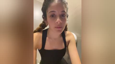 Media: Video of a young woman with light brown skin and braided hair, wearing a black tank top, looking directly at the camera with a neutral expression. Background shows tiled bathroom walls.