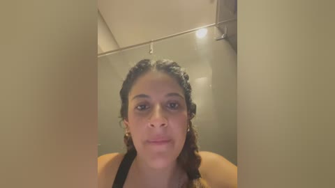 Media: A video of a young Black woman with light brown skin, braided hair, and a neutral expression. She is wearing a black top. The background features a beige wall and a ceiling light.