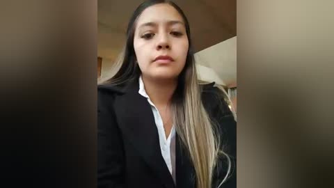 Media: A video of a young Asian woman with long, straight, dark brown hair and light skin, wearing a black blazer over a white shirt, standing in a dimly lit room with blurred background.