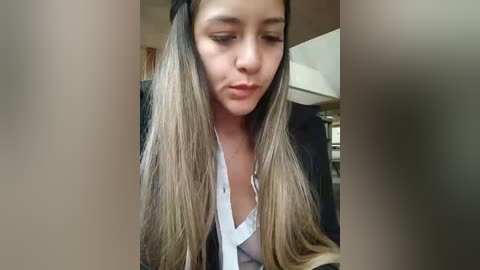 Media: Video of a young woman with long, straight, light brown hair, wearing a black blazer over a white shirt, making a pouty face, in a blurred indoor setting.