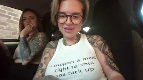 Media: Video of a tattooed woman in glasses wearing a white tank top with a provocative slogan inside a car, with another tattooed woman in the background.