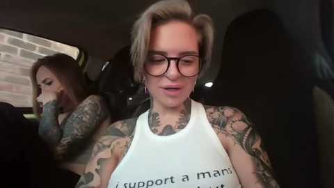 Media: Video of a tattooed woman with short blonde hair, wearing glasses and a white tank top with \"support a manatee\" text, sitting in a car.