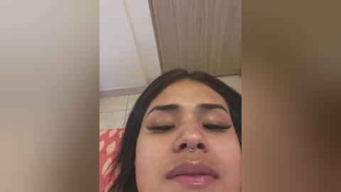 Media: Video of a young woman with medium brown skin, closed eyes, and a nose ring, lying on a red and white patterned rug. Background shows a beige wooden door and a white wall.