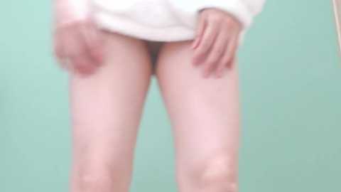 Media: Video of a light-skinned person from mid-thigh to upper torso, wearing a white sweater and no pants, standing against a green background, with hands on thighs.