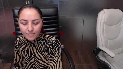 Media: Video of a woman with light skin and brown hair, wearing a zebra-patterned blouse, sitting on a black leather chair in an office with a beige chair and glass walls.