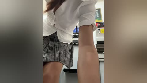 Media: Video of a woman in a white blouse and gray plaid skirt leaning over a desk, with blurred background featuring office equipment.