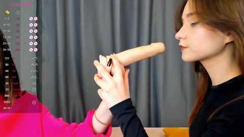 Media: A video shows a woman with long brown hair, wearing a black top, holding a large, light-colored dildo. She is focused on the dildo, which is nearly as tall as her. The background features gray curtains.