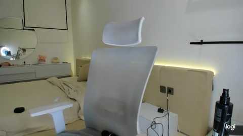 Media: Video of a minimalist bedroom with a modern white ergonomic chair, beige bed, white wall-mounted shelves, and a black bottle on the floor, illuminated by ambient lighting.