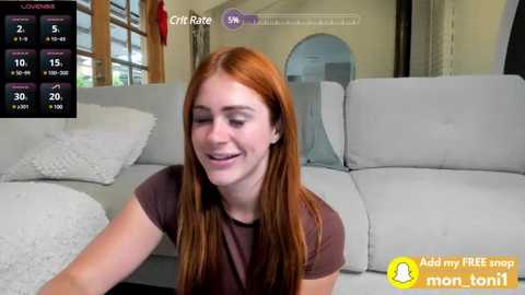 Media: Video of a fair-skinned, red-haired woman in a brown t-shirt, smiling, seated on a light gray couch in a modern living room with large windows and a white textured blanket.