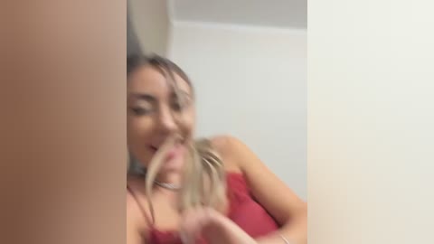 Media: A blurred video of a blonde woman with fair skin, wearing a red top, playfully biting the end of her hair. Background shows a white wall.