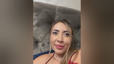 Media: Video of a smiling Latina woman with light skin, straight brown hair, and wearing a red lace top. She has a pearl necklace and sits on a grey tufted leather couch.