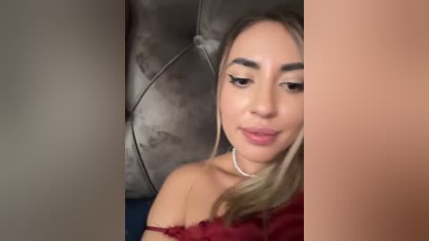 Media: Video of a young woman with light skin, blonde hair, and a red off-shoulder top, sitting on a gray, tufted leather couch.