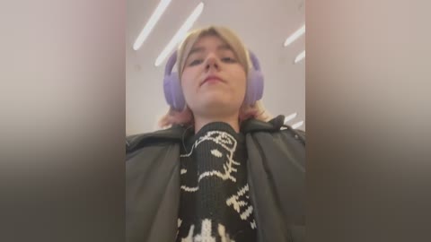 Media: A low-angle video of a young woman with light skin and blonde hair wearing purple headphones, a black jacket, and a sweater with white graphic designs, taken in a brightly lit, modern indoor setting.