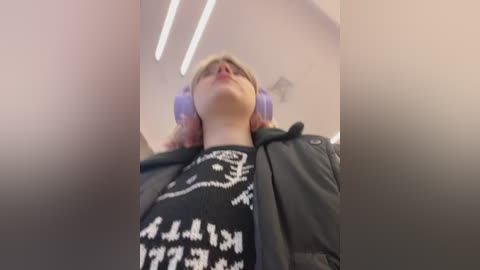 Media: Video of a young person with light skin and light brown hair, wearing purple headphones and a black jacket with a white graphic pattern, looking upward in a brightly lit, modern indoor setting.