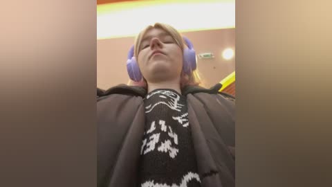 Media: Video of a young person with pale skin and light hair, wearing large purple headphones and a black sweater with white graphic patterns, standing indoors with blurred background.